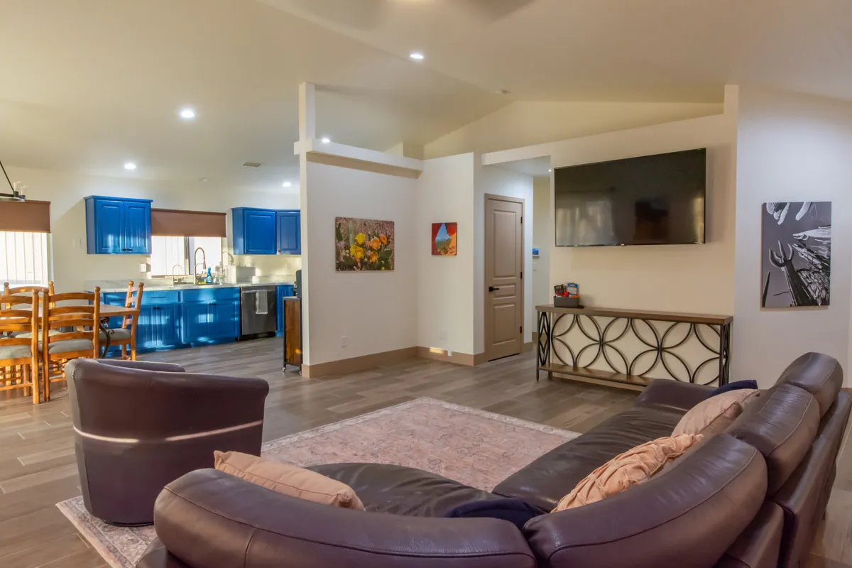 This image features a welcoming living room with comfortable leather sofa, blue kitchen cabinetry in the background, a dining space with wooden chairs, and decorative wall art. The space is brightened by natural light coming from large windows, and the furnishings set an inviting, cozy mood ideal for relaxation or gathering.