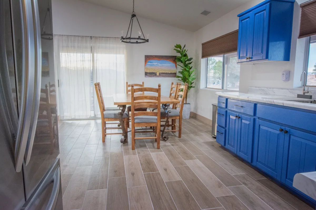 A bright and cheerful dining area with a wooden dining table, blue cabinetry, modern lighting, wall artwork, and an indoor plant. The setting feels inviting and conducive to group gatherings or intimate family meals.