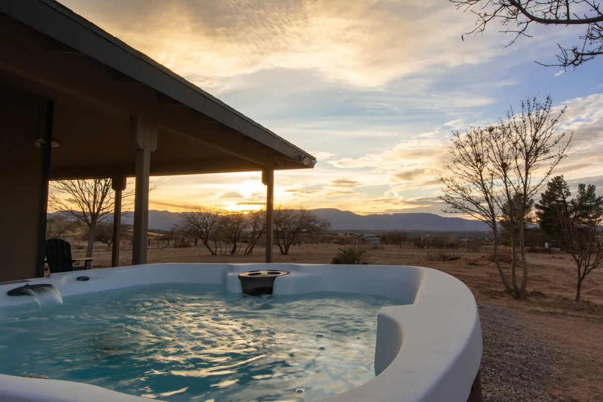 The image shows a spacious hot tub on a wraparound porch overlooking stunning Arizona mountains, with the vibrant colors of sunset glowing over the landscape. Tall trees frame the view, and the tranquil setting invites relaxation. The hot tub is steaming, hinting at soothing warmth, while a glass of wine rests nearby, evoking a sense of leisure and indulgence.