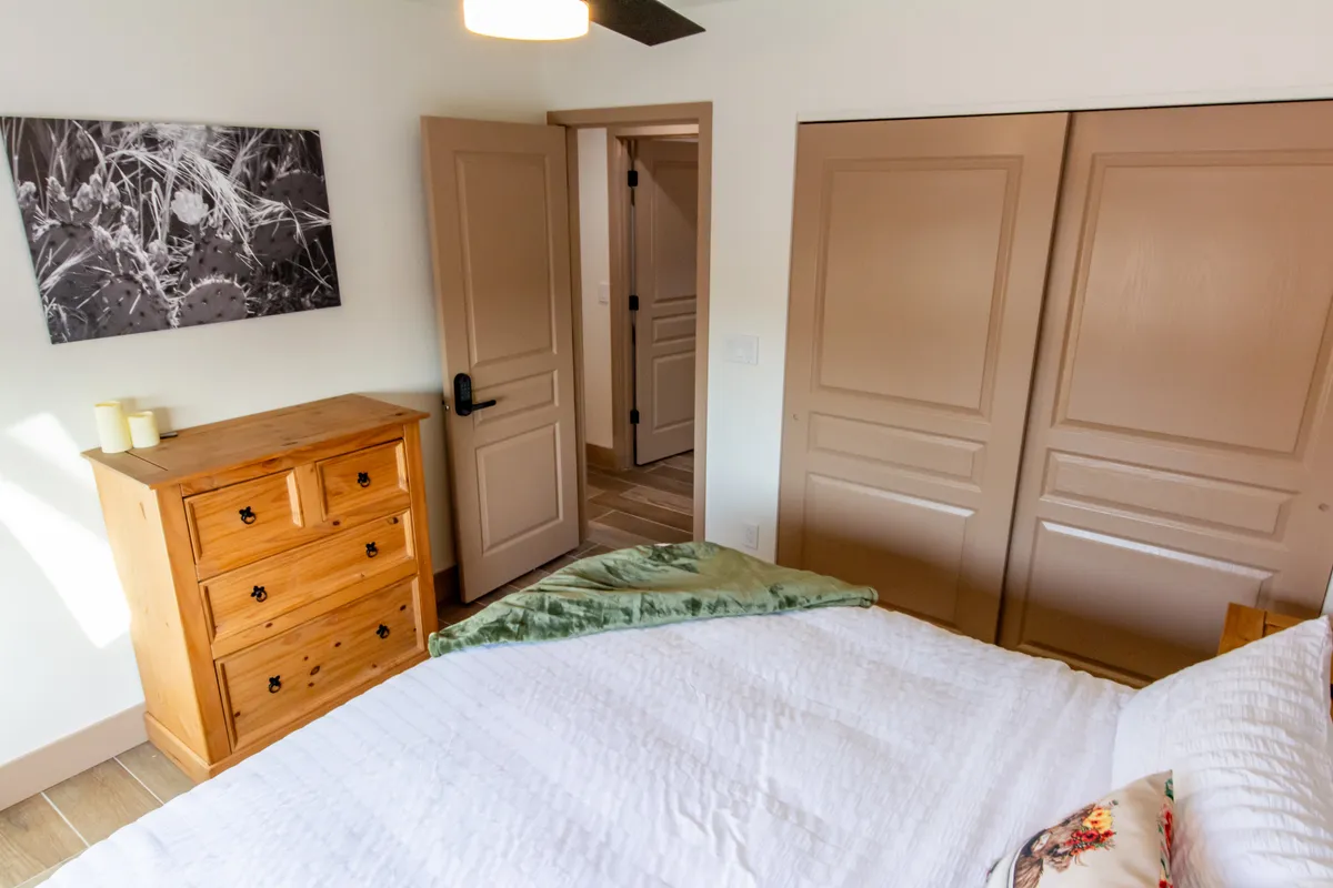 A cozy and tranquil interior bedroom scene featuring a wooden dresser, textured bedding, a green throw blanket, and artwork on the wall. Sunlight streams in, illuminating the restful space that invites relaxation and peaceful moments.
