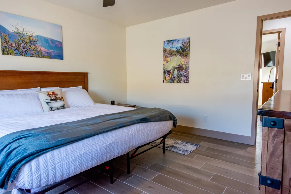 A tranquil bedroom with a large bed covered in crisp white linens, a decorative pillow, soft natural light, and serene landscape artwork above the headboard. A wooden dresser sits to the side, and the atmosphere feels restful and uncluttered—ideal for unwinding after a day of adventure or relaxation.
