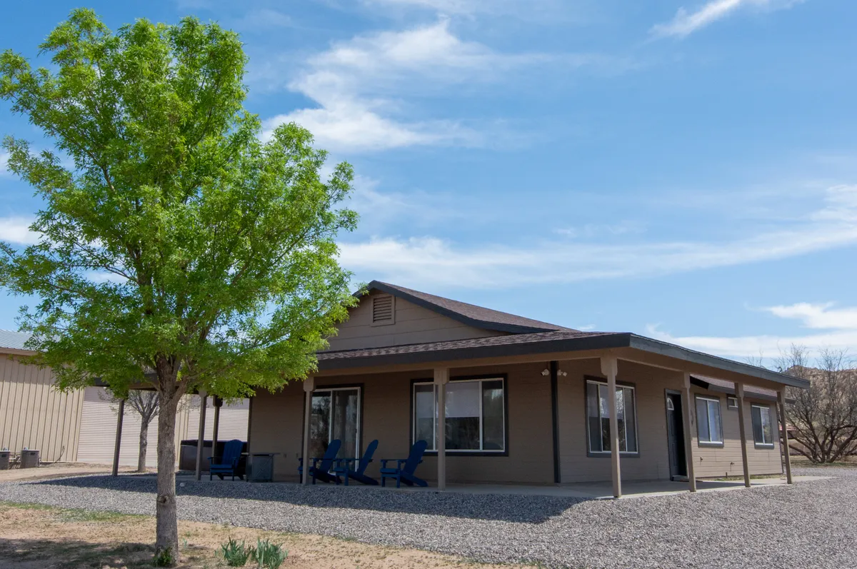 An exterior shot focusing on the wrap-around porch with chairs, a prominent tree providing shade, and large windows looking into the house. The atmosphere is serene, suggesting a seamless transition from indoors to outdoors, perfect for relaxing or stargazing as the sun sets.