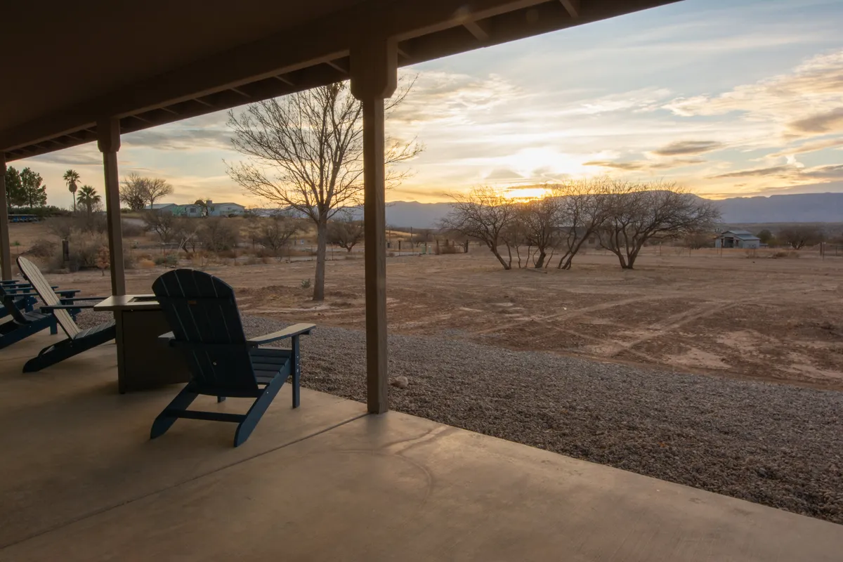 A cluster of adirondack chairs faces a dramatic sunset over distant mountains, with bare trees framing the serene, open landscape. The tranquil setting is ideal for stargazing, evening conversations, or quiet reflection in nature.