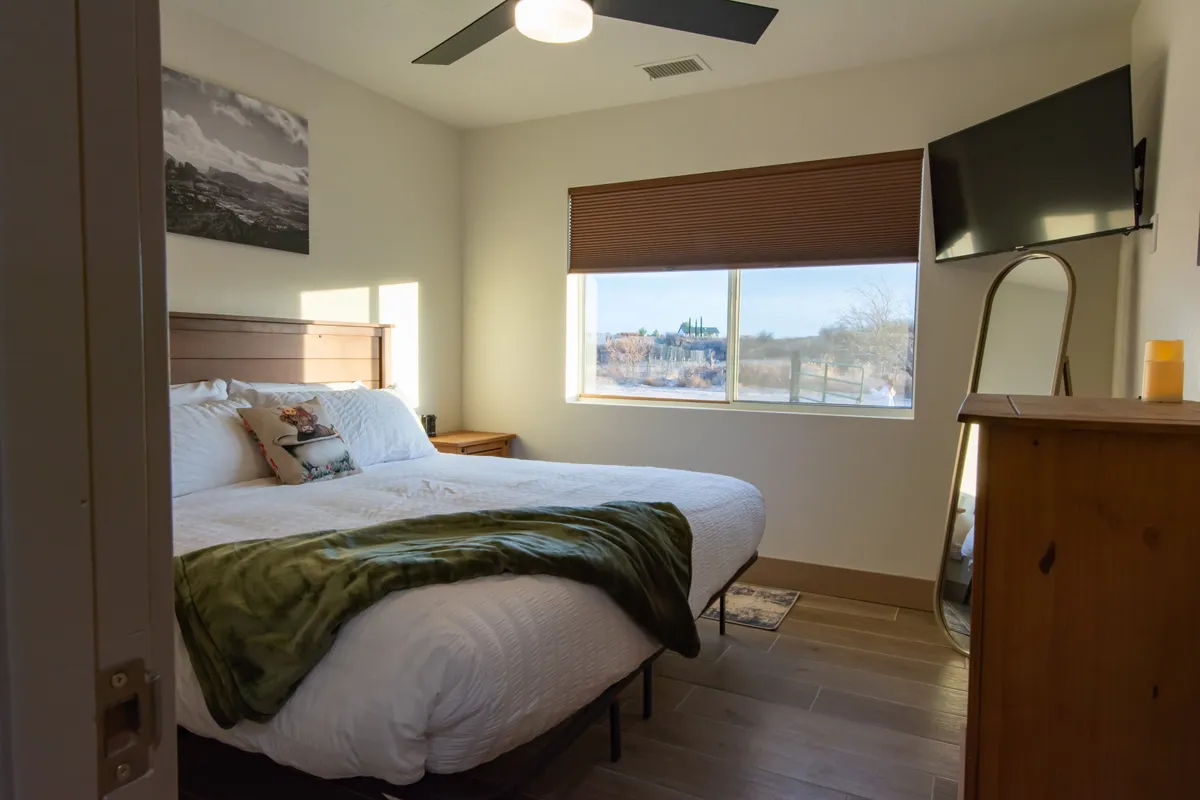 A welcoming king-sized bed with a decorative pillow faces a window letting in natural light. Beside the bed is a wooden dresser, and a TV is mounted on the wall, offering both comfort and entertainment. The soft glow and neat arrangement convey a cozy, restful ambiance.