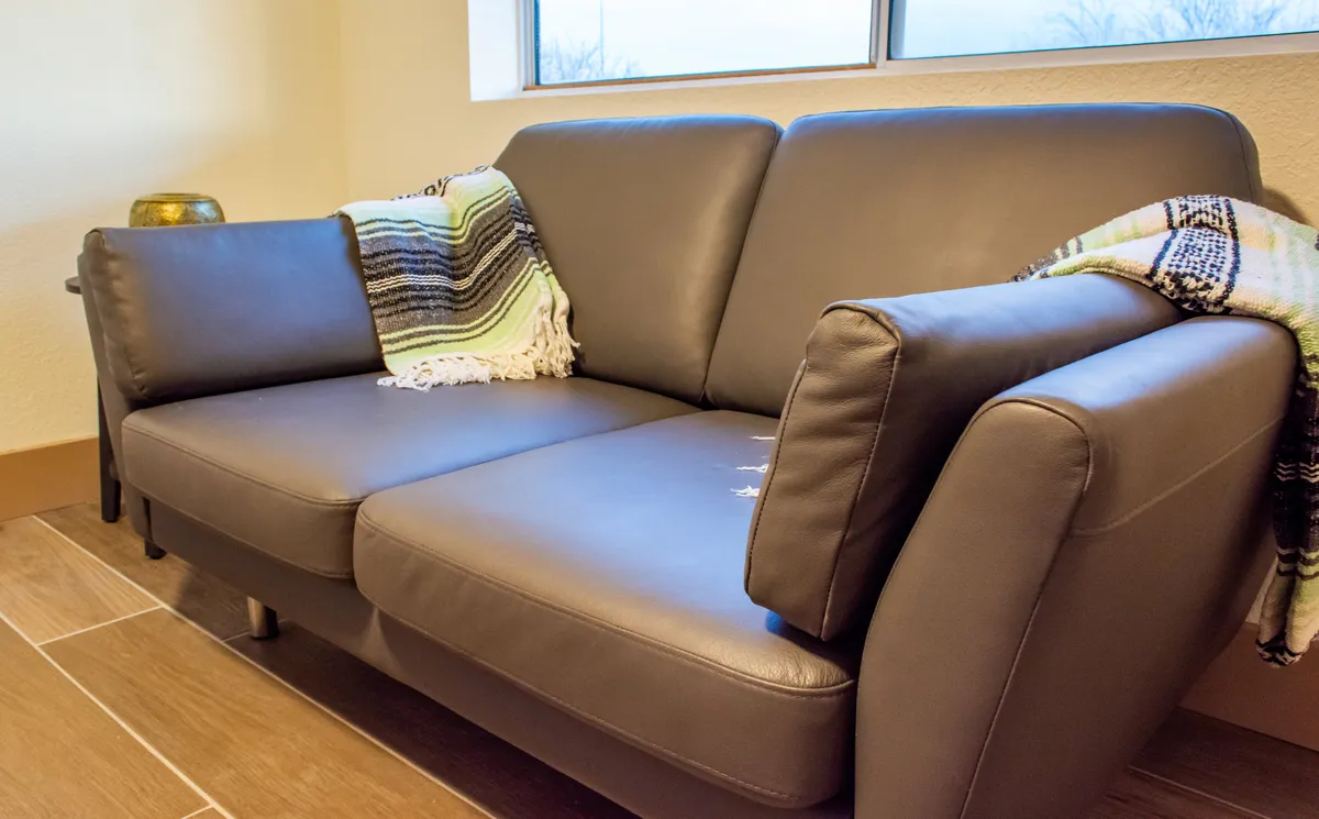 A bright, inviting living space featuring a modern sofa adorned with a textured throw blanket, positioned to soak in natural sunlight from a large window. The arrangement encourages relaxation, reading, or socializing and offers a glimpse of the outdoors through the window.