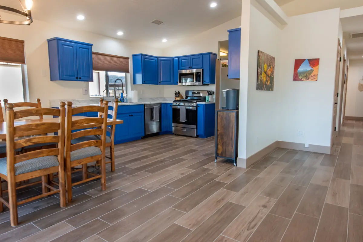 A warm, inviting kitchen scene featuring blue cabinets, a wooden dining table, and stainless steel appliances. The space is clearly designed for both cooking and gathering, with ample workspace and a dining area that encourages conversation and shared meals.