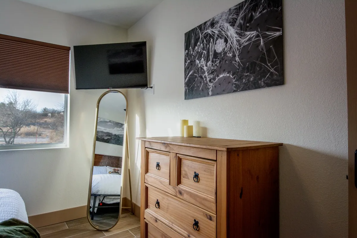 A modern bedroom scene featuring a large mirror, a wooden dresser, a TV, stylish black and white artwork, and a window streaming in soft morning light. The room feels inviting, contemporary, and perfectly suited for both relaxing and preparing for the day.